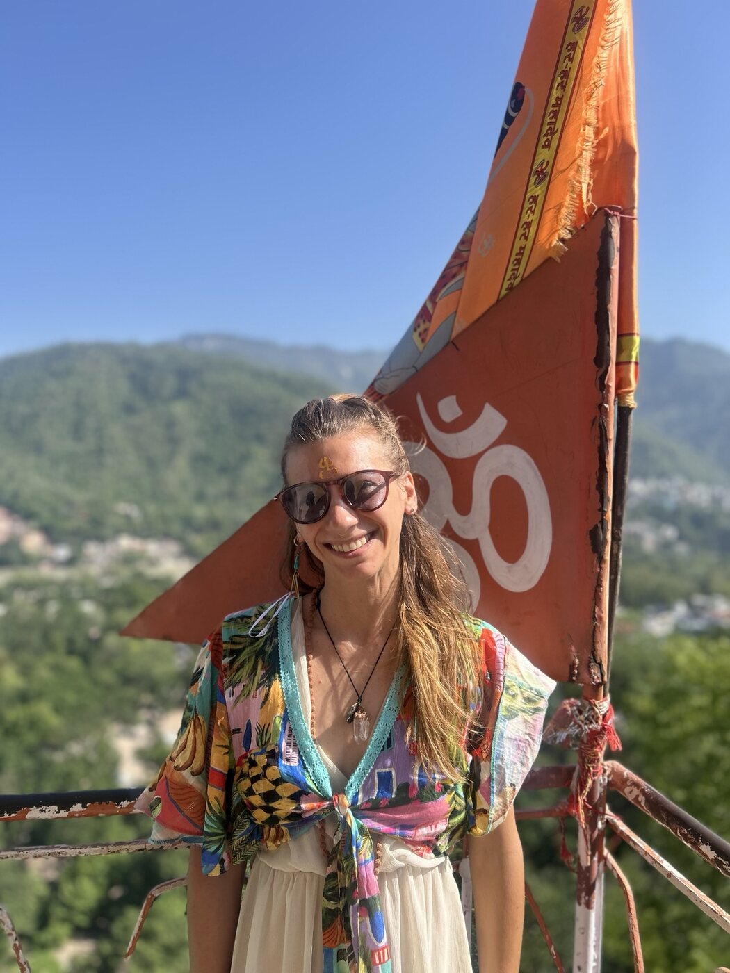Vanessa smiling beside an Om flag overlooking the hills of Rishikesh